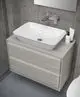 Waschtisch mit ovalem, weißem Becken auf einer hellen Holzkommode. Über dem Waschbecken befindet sich eine chromfarbene Armatur. Hintergrund mit grauer Wandtextur. Ideal Standard.